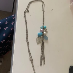 Halsband - Unikt långt halsband med silverfärgad kedja, dekorerat med blå, turkosa och lila pärlor samt ett stort silverfärgat blad. Hängande kedjor ger extra detalj. Perfekt för dig som gillar boho-vibe och färgglada accessoarer.❤️Aldrig använd! Ca 44cm långt. Perfekt som present eller gåva💕