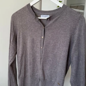 Grå kofta från Holly & Whyte - Snygg grå kofta från Holly & Whyte med rund halsringning och knappar framtill. Långärmad modell i mjukt material, perfekt att slänga över en t-shirt eller klänning. Stilren och enkel design som funkar till det mesta.