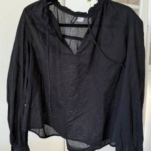 Svart blus med snörning från H&M - Snygg svart blus från H&M i tunn och luftig bomull. Blusen har v-ringning med snörning vid halsen och långa ärmar med resår vid handlederna. Perfekt för en chill och stilren look.