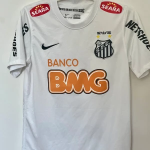 Santos Neymar Jr fotbollströja Nike - Vit Santos fotbollströja från Nike med Neymar Jr och nummer 11 på ryggen. Tröjan har korta ärmar, klubbmärke på bröstet, flera sponsorloggor och är tillverkad i lätt, andningsbart material. Perfekt för dig som älskar brasiliansk fotboll och Neymar.