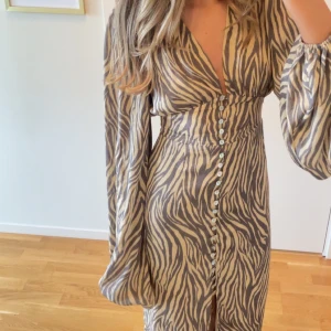 adoore klänning positano - Snygg långklänning med zebramönster i beige och brunt. Djup v-ringning, långa puffärmar och knappar längs hela framsidan. Klänningen har en markerad midja och slits framtill. Perfekt för dig som vill sticka ut med en trendig look.