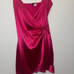 Rosa klänning i satin - Snygg kort omlottklänning i glansig rosa satin med knyt i sidan. Klänningen har v-ringning och är ärmlös, perfekt för dig som vill sticka ut med en färgstark look. Materialet är mjukt och följsamt med en lyxig känsla. Har två streck som jag befogat men inget direkt synligt!