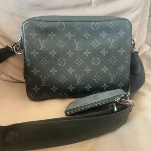Louis Vuitton svart axelväska monogram - Snygg svart axelväska från Louis Vuitton med klassiskt monogrammönster i grått. Väskan har bred axelrem med logga, två fack med dragkedja och en liten avtagbar plånbok. Tillverkad i skinn med silverfärgade metalldetaljer. Perfekt för dig som vill ha en stilren och lyxig look.
