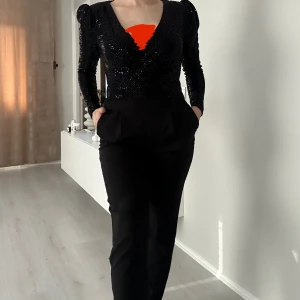 Svart Jumpsuit med paljetter - Snygg svart omlottjumpsuit med v-ringning och långa ärmar. Jumpsuiten är täckt av små paljetter som ger en glittrig, iögonfallande look och har markerade axlar för en trendig siluett. Perfekt för dig som vill sticka ut med en cool och festlig vibe – allt i ett enda plagg!
