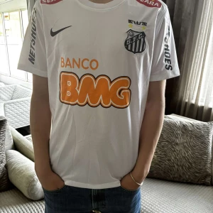 Vit Santos Neymar Jr fotbollströja Nike - Santos FC matchtröja i vitt med Neymar Jr och nummer 11 på ryggen. Tröjan har svarta tryck på ärmarna, klubbmärke på bröstet och stora orange BMG-loggor fram och bak. Tillverkad i lätt och ventilerande material, klassisk passform och rund hals.