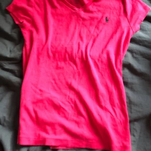 Rosa t-shirt från Polo Ralph Lauren - Snygg rosa t-shirt från Polo Ralph Lauren med klassisk v-ringning och korta ärmar. T-shirten har det ikoniska svarta logobroderiet på bröstet och är gjord i mjuk bomull. Perfekt för en chill och stilren look. Tröjan har en liten fläck som vid urringningen men det är inget som märks på avstånd.  