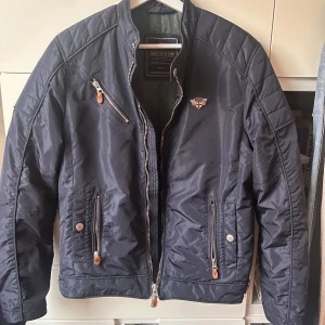 Morris marinblå bomberjacka S - Snygg marinblå bomberjacka från Morris med coolt emblem på bröstet och flera dragkedjeförsedda fickor. Jackan har quiltade detaljer och är tillverkad i glansig polyester. Perfekt för dig som vill ha en stilren och sportig look.