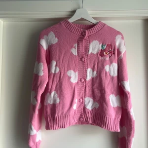 Care Bears x Skinnydip rosa kofta - Supergullig limited edition rosa kofta med vita moln från Care Bears x Skinnydip. Broderad Care Bear med vingar och pilbåge på bröstet, texten 'angel' under. Koftan är stickad, har långa ärmar och knäpps med matchande knappar framtill.