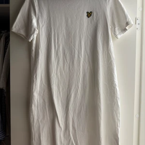 Vit t-shirtklänning från Lyle & Scott strl M - Vit t-shirtklänning från Lyle & Scott med klassisk passform och korta ärmar. Klänningen har en enkel design med det ikoniska gula fågelmärket broderat på bröstet. Tillverkad i mjuk bomull för en skön känsla hela dagen. Aldrig använd. 