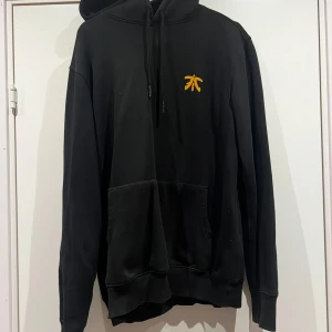 Svart Fnatic hoodie XL - Svart hoodie från Fnatic i storlek XL med orange Fnatic-logga på bröstet och stor text 'ALWAYS FNATIC' på ryggen. Klassisk känguruficka, huva med snörning och mjukt material. Perfekt för dig som älskar e-sport och vill ha en stilren streetwear-look.