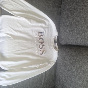 Vit sweatshirt från Hugo Boss - Vit sweatshirt från Hugo Boss med broderad logga i stora bokstäver framtill. Tröjan har rund halsringning och långa ärmar. Tillverkad i mjuk bomull, perfekt för en clean och stilren look.