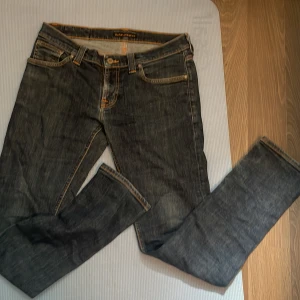 Mörkblå jeans från Nudie Jeans - Säljer ett par mörkblå jeans från Nudie Jeans med klassisk orange kontrastsöm och snygga detaljer på bakfickorna. Jeansen har rak passform och fem fickor. Perfekta för dig som gillar stilrena och tidlösa jeans.