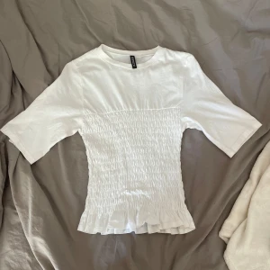 Vit smockad topp med volangkant - Supersöt vit topp från Monki med smockad midja och volangkant nertill. T-shirtrmodell med korta ärmar och rund halsringning. Perfekt för dig som gillar en feminin och trendig stil.