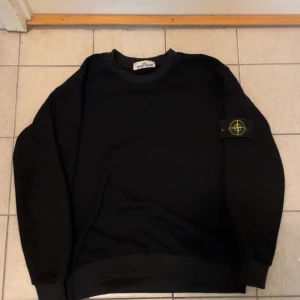 Svart Stone Island sweatshirt - Svart sweatshirt från Stone Island med klassisk logga på vänster ärm. Tröjan har rund halsringning, ribbade muddar och är långärmad. Perfekt för dig som gillar stilrena och ikoniska plagg med streetkänsla.