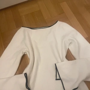 Vit långärmad topp med svarta kanter - Vit långärmad topp med svarta kanter vid halsringning och ärmslut. Ärmarna har en unik volangdetalj som ger en cool och trendig look. Perfekt för dig som gillar stilrena plagg med en twist. Materialet känns mjukt och skönt mot huden.