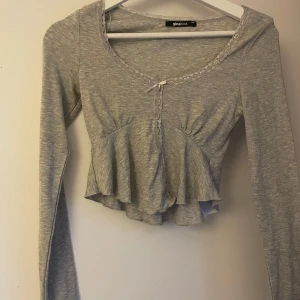 Grå volangblus med spets från Gina Tricot - Supersöt ljusgrå blus från Gina Tricot med volang nertill, långa ärmar och spetsdetaljer längs ringningen och framtill. Liten rosett framtill ger extra gullig touch. Perfekt för dig som gillar romantisk och trendig stil.