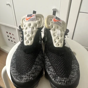 Nike x Off-White Vapormax sneakers - Svarta och vita Nike x Off-White Vapormax sneakers med stickad ovandel, tydliga Off-White detaljer och text på sidan. Skorna har en unik design med vit plös, svarta snören och synlig sula. Perfekta för dig som gillar streetwear och vill sticka ut.