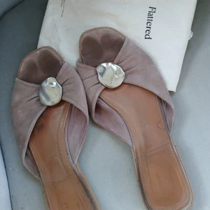 sandaler från Flattered strl 41 - Snygga sandaler från Flattered i beige mocka med breda band och en stor, oregelbunden silverfärgad metallknapp framtill. Platt sula och öppen tå ger en avslappnad men chic vibe. Perfekta för varma dagar när du vill ha något både stilrent och bekvämt.           100% mocka/läder. Använd skick. Dustbag medföljer 