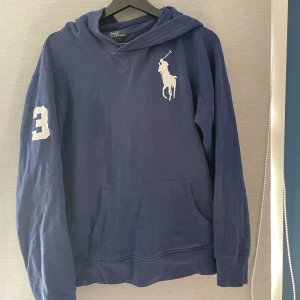 Marinblå hoodie Polo Ralph Lauren XL - Marinblå hoodie från Polo Ralph Lauren i storlek XL men passar M dock så är den lite bredare men inget som stör jätte mycket vid användning . Klassisk stor vit logga på bröstet och siffra 3 på ärmen. Tröjan har huva och en stor magficka framtill. Perfekt för dig som gillar sportig och avslappnad stil.