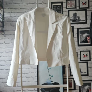 Vit croppad kavaj från Amisu - Snygg vit kavaj från Amisu i croppad modell med klassisk krage och en knapp framtill. Perfekt för dig som vill ha en stilren och fräsch look. Långärmad och med enkel bröstficka för extra detalj. Passar till både jeans och kjol. Använd till student har en fläck på ena axeln.