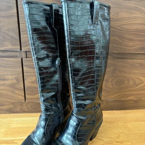 Svarta knähöga cowboyboots - Snygga svarta boots i croc-mönstrat syntetmaterial med spetsig tå och markerad klack. Höga skaft och dragkedja på insidan! Sparsamt använda! 