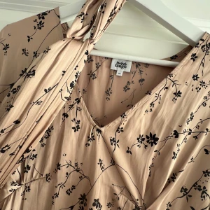 Beige blommig klänning Twist & Tango - Superfin beige klänning från Twist & Tango med svart blommigt mönster, v-ringning och knytband i midjan. Klänningen har vida trekvartsärmar och är tillverkad i återvunnen polyester. Perfekt för en somrig och avslappnad look.
