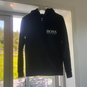Svart hoodie från Hugo Boss - Svart hoodie från Hugo Boss med vit logga på bröstet. Klassisk modell med huva, dragkedja och långa ärmar. Perfekt för en clean och stilren look. Mjuk bomullskänsla och normal passform. Denna tröja saknar neck tagen.