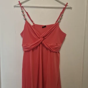 Korallrosa kortklänning med pärldetaljer - Supersöt rosa linne med twistad omlott framtill och tunna axelband dekorerade med silvriga pärlor. Linnet har en luftig passform och är perfekt för varma dagar.