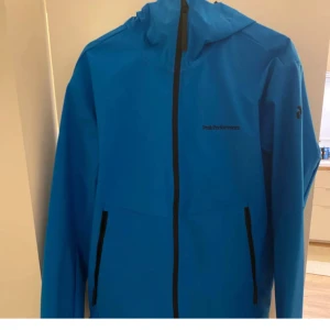 Blå vindjacka Peak Performance L - Snygg blå vindjacka från Peak Performance med svarta dragkedjor och huva. Jackan har en clean look och är perfekt för blåsiga dagar. Lätt och smidig att bära, med två praktiska fickor framtill och diskret logga på bröstet.