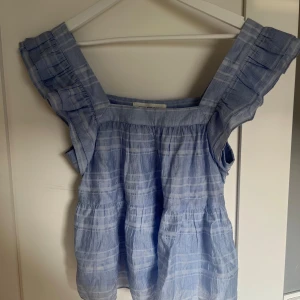 Blå rutig blus från Sofie Schnoor - Supersöt blå blus från Sofie Schnoor med rutigt mönster och volangdetaljer på axlarna. Blusen har fyrkantig ringning, är ärmlös och har en luftig, flowy passform. 