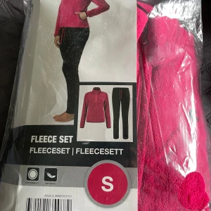 Rosa fleecejacka med dragkedja S - Mysig rosa fleecejacka i mjuk polyester med hel dragkedja framtill och hög krage. Jackan har svarta detaljer och en enkel, sportig look. Perfekt för kyliga dagar eller som ett extra lager när du vill vara bekväm.