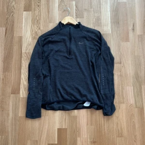 Nike Dri-Fit svart långärmad träningströja - Svart långärmad träningströja från Nike med Dri-Fit-teknologi. Tröjan har half zip framtill, diskreta reflexdetaljer på ärmar och rygg samt en sportig, slim passform. Perfekt för löpning eller gym. Materialet är syntet och andas bra.