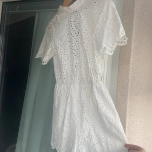 Vit jumpsuit med broderade detaljer - Superfin vit jumpsuit med broderade hålmönster och v-ringning i ryggen. Den har korta ärmar och resår i midjan för en snygg passform. Perfekt för sommarens alla tillfällen!