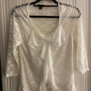 Vit spetsblus med stor rosett - Superfin vit spetsblus med trekvartsärm och en stor, elegant rosett framtill. Blusen är helt i spets med transparent känsla och har en feminin, romantisk vibe. Perfekt för dig som gillar detaljer och vill sticka ut lite extra.