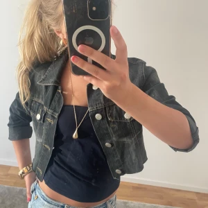 Svart croppad jeansjacka - Säljer en svart croppad jeansjacka med silvriga knappar och klassisk krage. Jackan har två bröstfickor med lock och långa ärmar. 