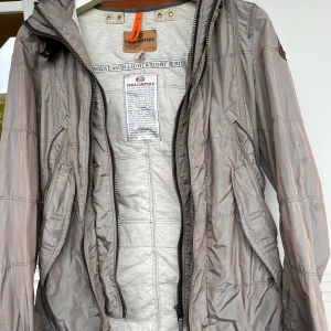 Grå vindjacka från Parajumpers S - Snygg grå vindjacka från Parajumpers med huva, dragkedja och coola detaljer som patch på ärmen och orange ögla i nacken. Jackan är lätt och perfekt för vår och höst. Flera fickor och justerbara detaljer bak för extra passform.