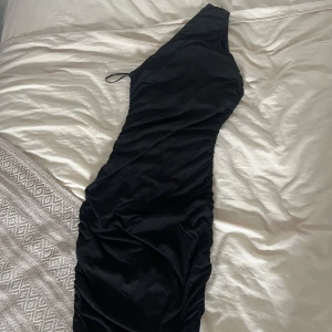 Svart långklänning one shoulder - Snygg svart långklänning med one shoulder-design och lätt rynkade detaljer längs sidorna. Klänningen har en tight passform och ärmlös stil med en axelbandslös look på ena sidan. Perfekt för dig som vill sticka ut med en elegant och modern siluett.