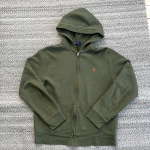 Polo Ralph Lauren hoodie  - Säljer en snygg olivgrön hoodie från Polo Ralph Lauren med dragkedja och det klassiska röda logotypbroderiet på bröstet. Perfekt för en avslappnad stil med långa ärmar och en bekväm huva. Passar perfekt för kyligare dagar. 🧥