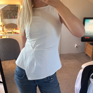 Vit ärmlös blus med asymmetrisk skärning - Säljer en stilren vit blus med ärmlös design och snygg asymmetrisk skärning framtill. Toppen har en lätt utsvängd nederdel som ger en modern siluett. Perfekt att matcha med jeans eller kjol för en clean look.