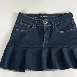 Mörkblå jeanskjol med volang Levi's - Snygg mörkblå jeanskjol från Levi's med volang nertill och rå kant. Klassisk femficksmodell med synliga sömmar och knappstängning framtill. 