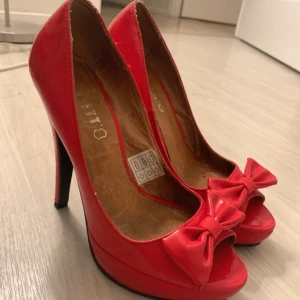 Röda pumps med rosett och öppen tå - Snygga röda pumps från Scorett med hög klack och plattform. Skorna har öppen tå och en söt rosett framtill. Tillverkade i glansigt syntetmaterial som ger en riktigt poppig look. Äkta läder på insidan, strl 37