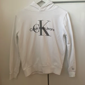 Vit hoodie från Calvin Klein Jeans - Snygg vit hoodie från Calvin Klein Jeans. Tröjan har huva, långa ärmar och ribbade muddar. Barnstorlek st 14 men passar en Xs.