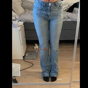Blå bootcut jeans - Snygga blå jeans med bootcut passform och medelhög midja. Jeansen har en slitning vid ena knät för en trendig look och klassiska fem fickor. Tillverkade i jeansmaterial med dragkedja och knapp framtill.