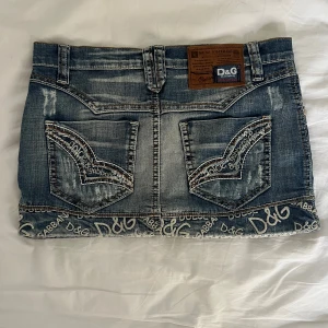 D&G jeanskjol med logga och slitningar - Snygg kort jeanskjol från Dolce & Gabbana med slitna detaljer och tydlig D&G-logga längs nederkanten. Klassisk blå denim med kontrastsömmar, bakfickor med broderad logga och läderpatch i midjan. Perfekt för dig som vill sticka ut med en ikonisk streetstil.