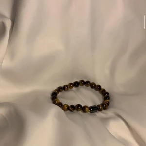 Brunt pärlarmband med tigeröga - Snyggt armband med runda pärlor i tigeröga-sten som skiftar i brunt och gult. Armbandet har elastisk tråd och en svart detaljpärla som accent. Perfekt accessoar för att lyfta din stil med naturliga vibes.