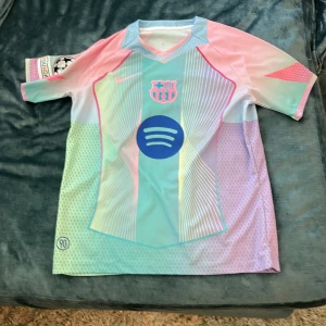 FC Barcelona Lamine Yamal Pastelltröja - Säljer en FC Barcelona fotbollströja från Nike med Lamine Yamal och nummer 10 på ryggen. Tröjan har korta ärmar, pastelliga färger i blått, rosa och mintgrönt, samt tryckta detaljer som Spotify-logga och UEFA Foundation-märke. Materialet är lätt och andas, perfekt för match eller träning.