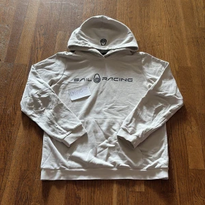 Vit hoodie från Sail Racing - Vit hoodie från Sail Racing med svart logga och tryck framtill. Klassisk huva och magficka, perfekt för en avslappnad stil. Mjuk och skön bomullskänsla, passar dig som gillar sportiga märken.
