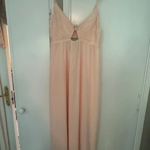 Ljusrosa långklänning med v-ringning - Superfin ljusrosa långklänning med smala axelband och v-ringad topp. Klänningen har ett snyggt veckat liv, liten cut-out framtill och faller mjukt i ett tunt, flowy tyg. Perfekt för sommarens speciella tillfällen!