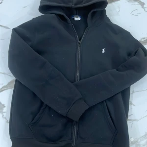 Svart hoodie från Polo Ralph Lauren - Svart hoodie från Polo Ralph Lauren med dragkedja framtill och klassisk huva. Tröjan har lång ärm, ribbade muddar och en liten vit broderad logga på bröstet. Perfekt för en chill och stilren look.