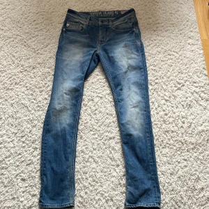 Blå jeans från Garcia Jeans - Säljer ett par klassiska blå jeans från Garcia Jeans med snyggt slitna detaljer och femficksmodell. Jeansen har normal passform och raka ben, tillverkade i bomull med en autentisk tvättad look. Perfekta för en avslappnad och cool stil.